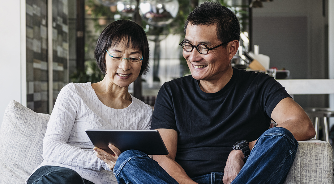 Couple sitting on a couch, wearing glasses, looking at a tablet, and smiling