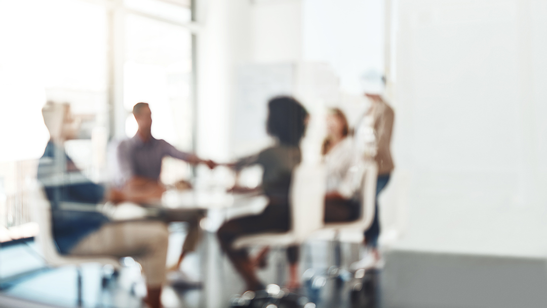 Out of focus image of a team of five people meeting around a table, two are shaking hands across the table